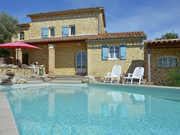 A stone building with a swimming pool, outdoor seating, and a red umbrella.