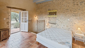 A bedroom with a stone wall, a bed, a nightstand, a lamp, and a door leading to an outdoor area. - Villa in Les Mages with swimming pool (Les Mages)