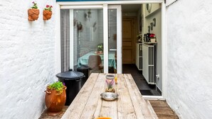 Une table en bois, un bac à ordures et des plantes en pot se trouvent dans un espace extérieur aux murs blancs, avec une porte-fenêtre coulissante donnant sur un espace intérieur.