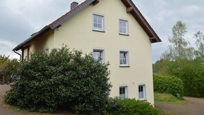 A two-story house with a sloped roof, multiple windows, and a well-maintained garden with bushes and a tree. - Comfy Apartment in Gransdorf with Garden (Gransdorf)
