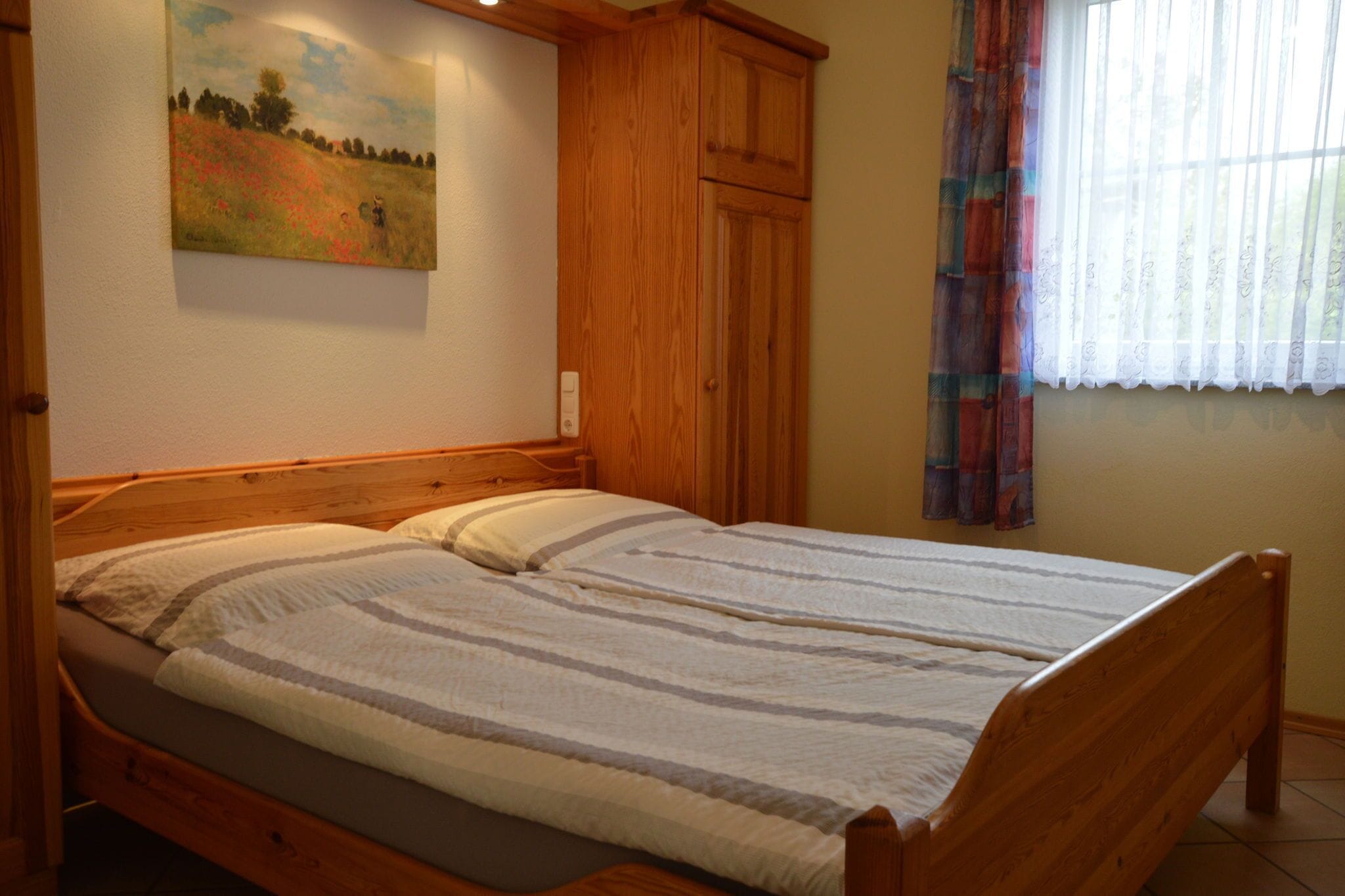 Una cama doble con estructura de madera y colcha a rayas, un armario de madera y un cuadro de un campo en la pared.