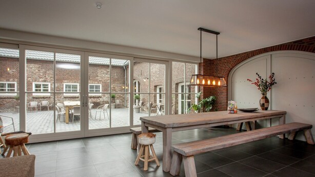 A spacious indoor area with a large sliding glass door leading to an outdoor patio. There is a wooden table with benches, a hanging light fixture, and a vase with flowers on a side table. - Pet-Friendly Mansion (Swolgen)
