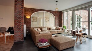 A modern living room with a sectional sofa, ottoman, and coffee table. There is a large arched window and a brick wall. - Pet-Friendly Mansion (Swolgen)