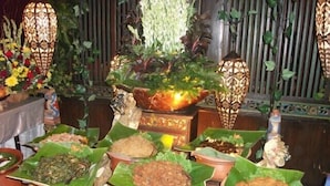 Presentación tradicional de comida con alimentos servidos en hojas de plátano en vasijas de barro, rodeado de faroles decorativos y follaje.