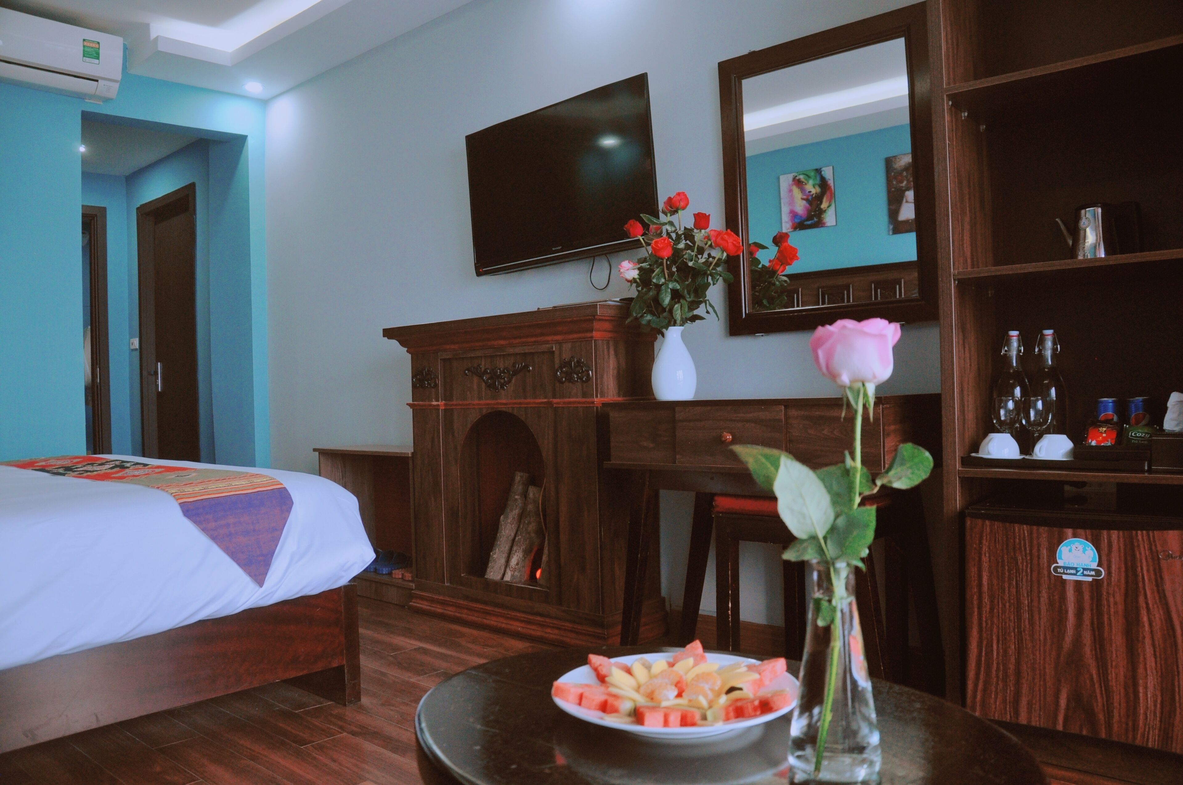 Une chambre d’hôtel avec un lit, un bureau, un vase de fleurs, une assiette de fruits et un téléviseur fixé au mur.