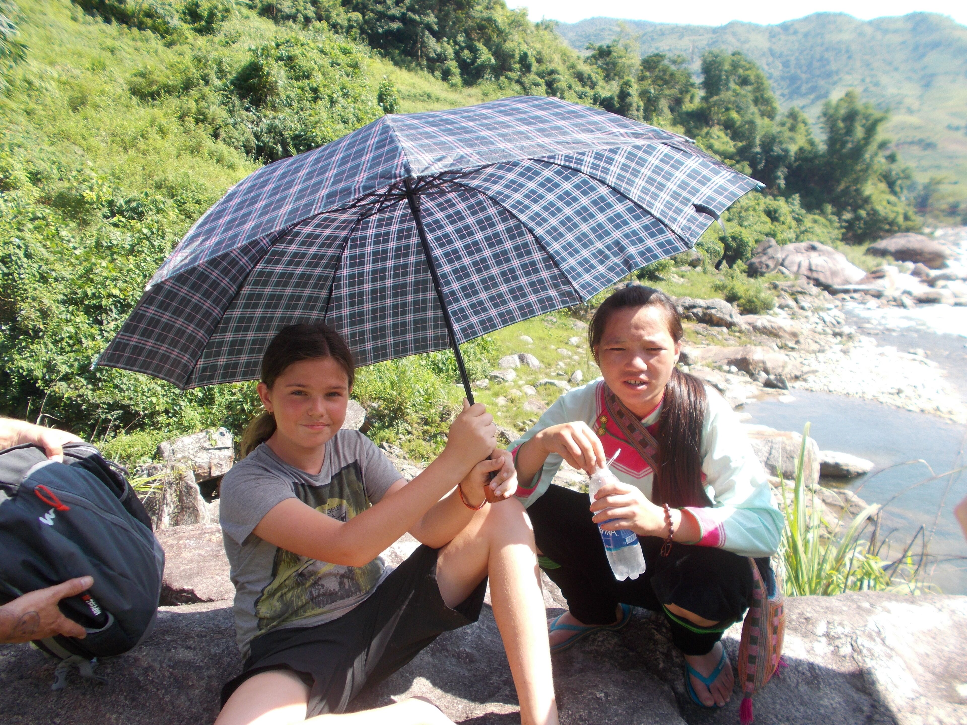Two people sitting on rocks by a river, one holding a plaid umbrella.