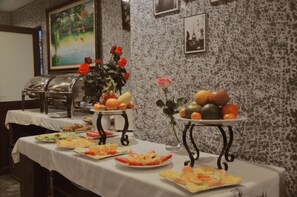 Una mesa de buffet con frutas y verduras variadas, un florero decorativo con rosas rojas y un cuadro en la pared.
