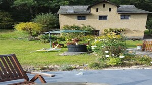 A wooden deck with a glass table and chairs, a stone fire pit, and a house with a garden in the background.