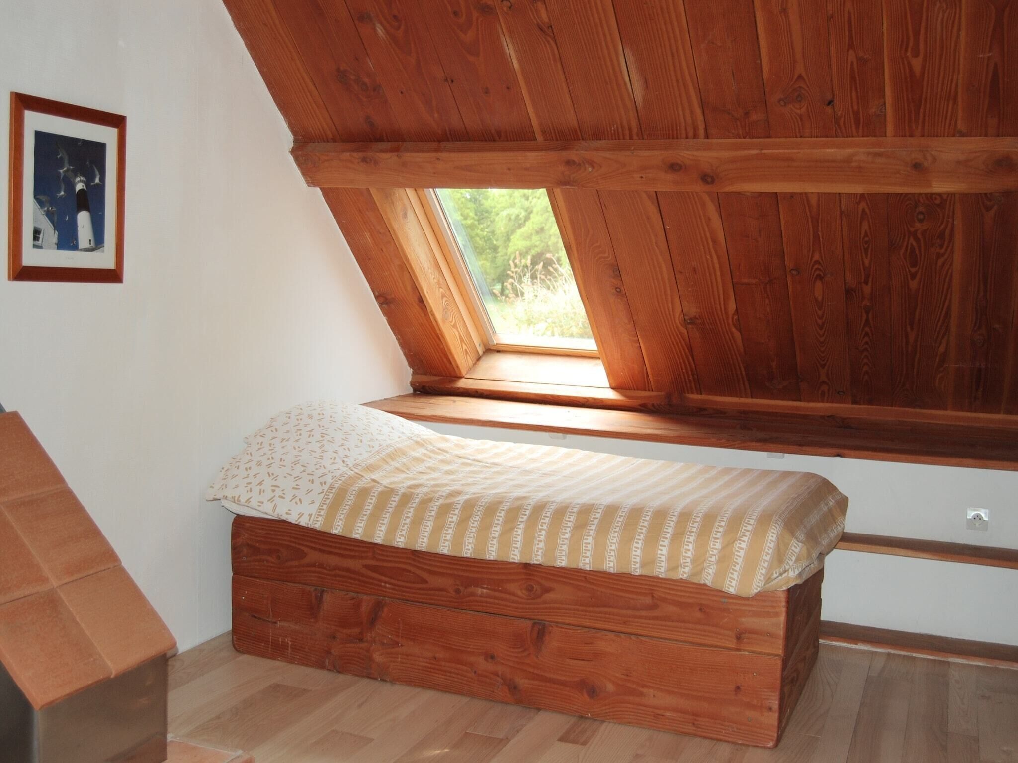 Ein Schlafzimmer im Loft-Stil mit schrägverlaufender Decke, einem Fenster, einem Bett mit gemustertem Überwurf und einem gerahmten Bild an der Wand.