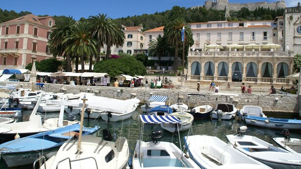 Un port de plaisance avec des bateaux amarrés, un bâtiment historique doté d’arcades et, au loin, sur une colline, un château.