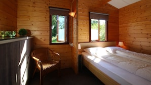 Ein Zimmer in einer Holzhütte mit einem Bett, einem Sessel, einem kleinen Tisch und zwei Fenstern.