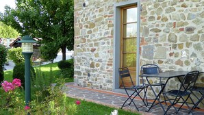 A stone building with a wooden door and a window, a small outdoor table and chairs, and a garden with flowers and shrubs. - Holiday Home in Montecarelli with Pool (Montecarelli)