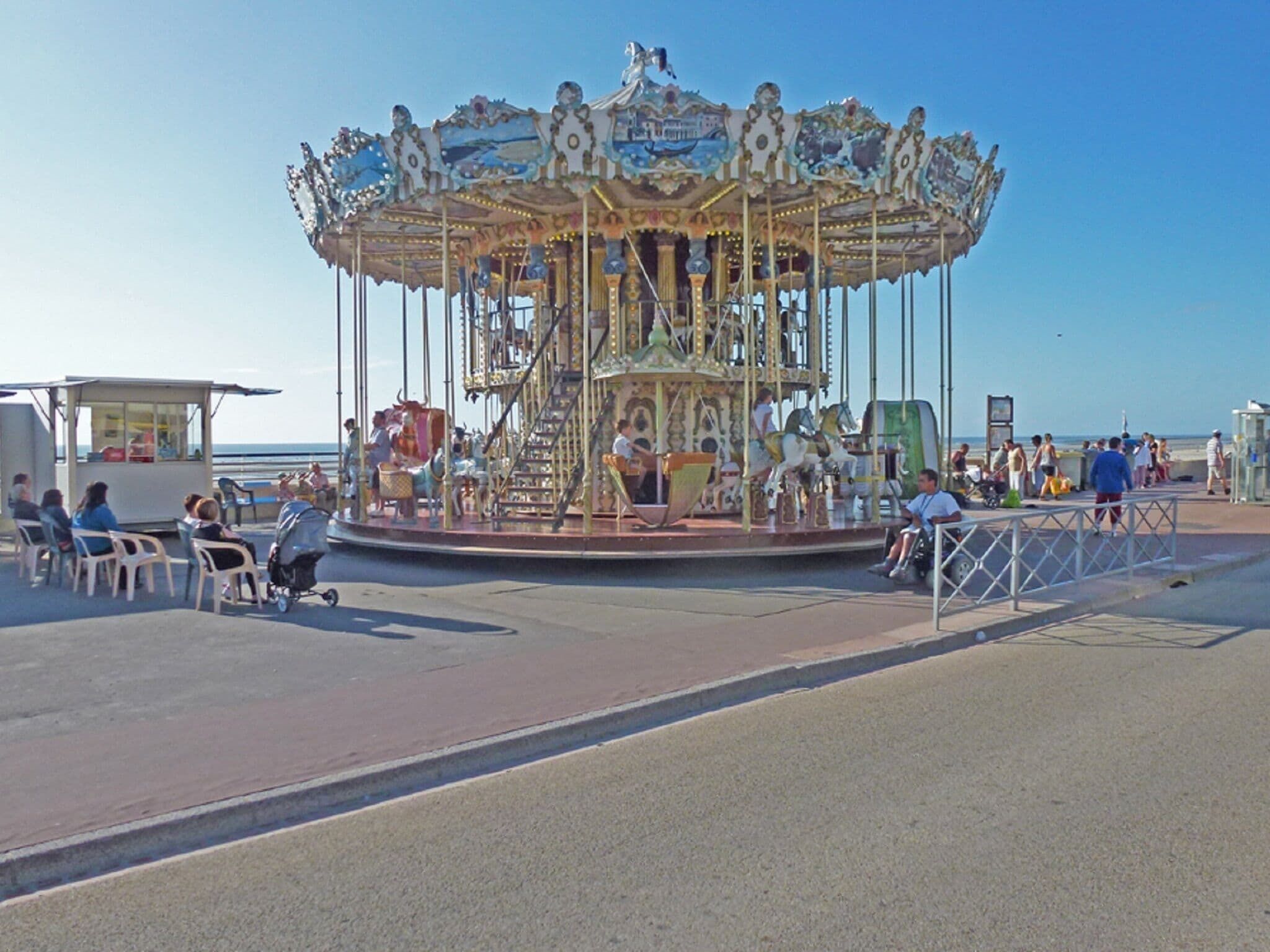 A carousel with ornate decorations and horses, surrounded by a paved area with chairs and a railing.