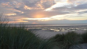 Ein Strand mit Dünen und Gras im Vordergrund, und ein Sonnenuntergang über dem Meer.