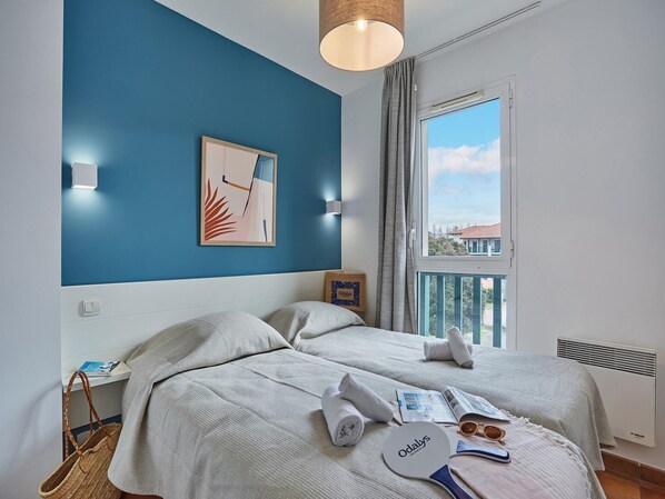 A hotel room with two beds, a window with a view, a painting on the wall, and a towel and sunglasses on the bed.