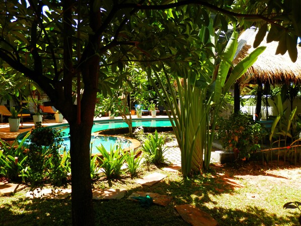 Una piscina rodeada de exuberante vegetación, una estructura con techo de paja y un jardín bien cuidado.