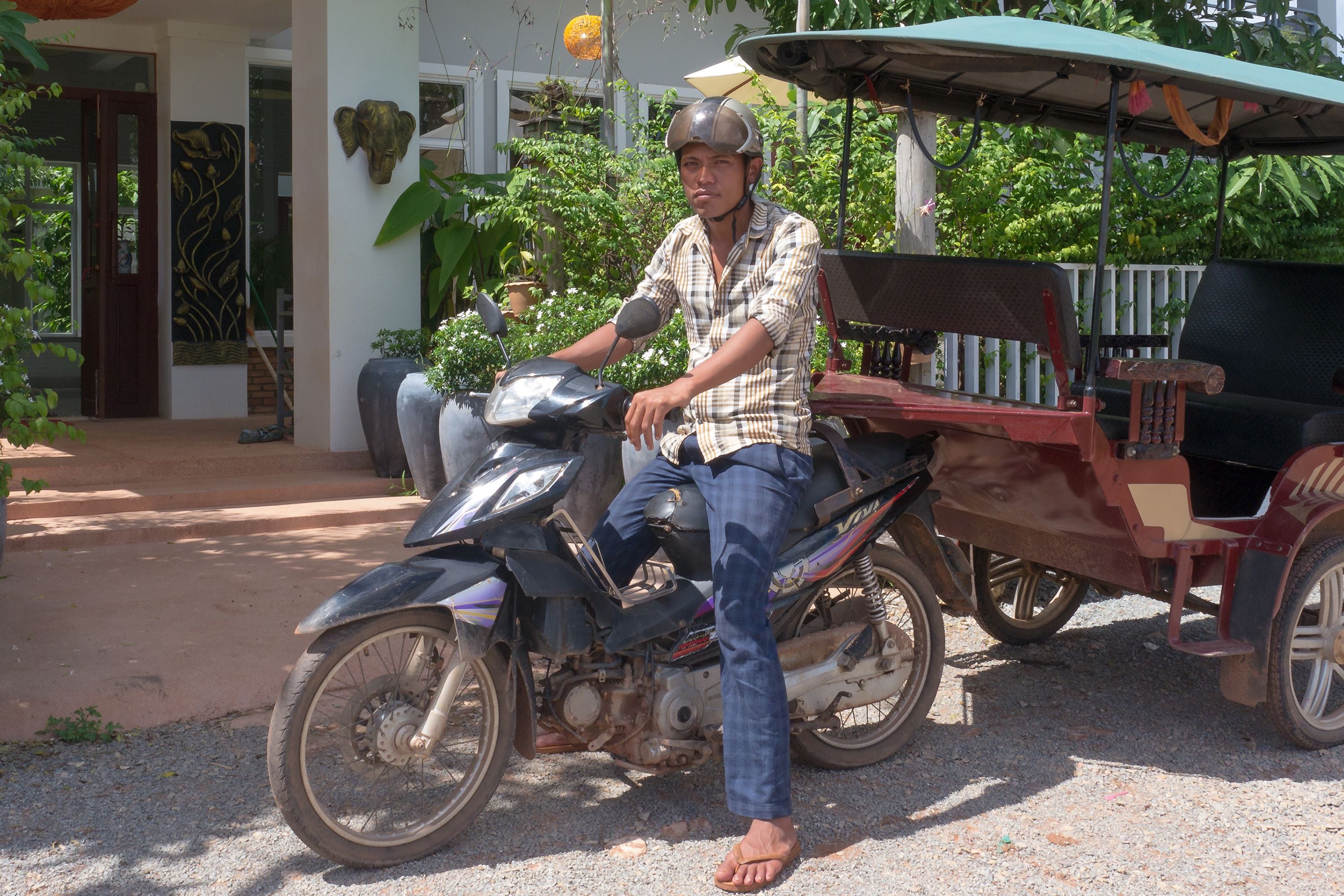A man sitting on a scooter parked in front of a building with a porch and potted plants.