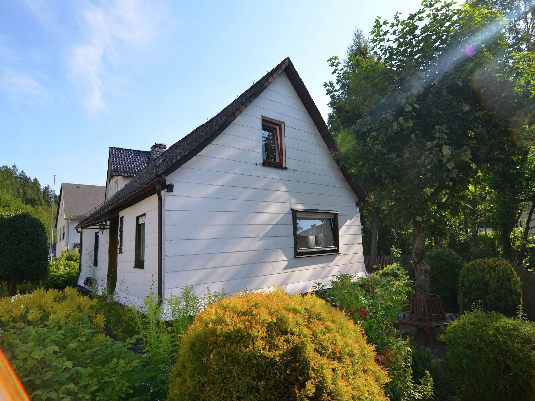 Une maison blanche avec un toit à pignon, entourée d’une végétation luxuriante et d’un jardin bien entretenu.