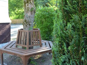 A wooden bench with a backrest and armrests, positioned on a gravel path beside a tree and dense green foliage. - Holiday home in Ramsbeck with garden (Bestwig-Ramsbeck)