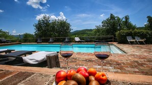 A poolside setting with a bowl of fruit and two glasses of red wine on a brick surface.
