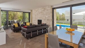 A modern living room with a fireplace, a dining area with a glass table, and a view of a swimming pool.