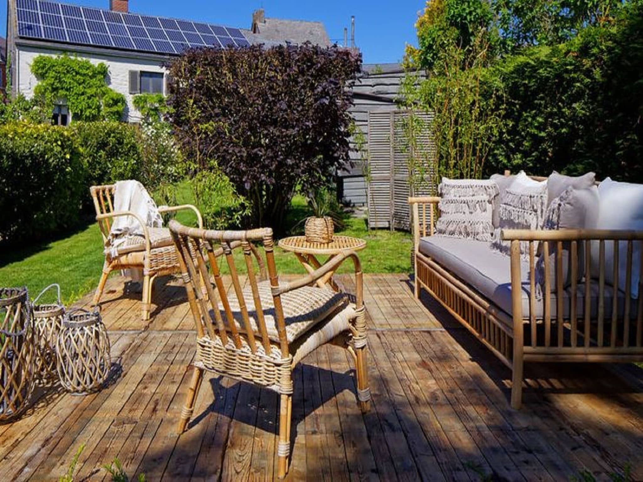 Une terrasse en bois, agrémentée de meubles en osier, dont des chaises et un canapé, entourée de verdure et bordée d’une maison dotée de panneaux solaires.