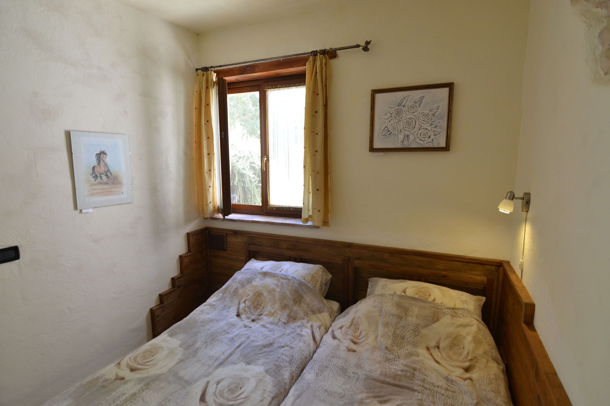 A bedroom with a wooden headboard, two beds with patterned bedding, a window with curtains, and two framed pictures on the wall.