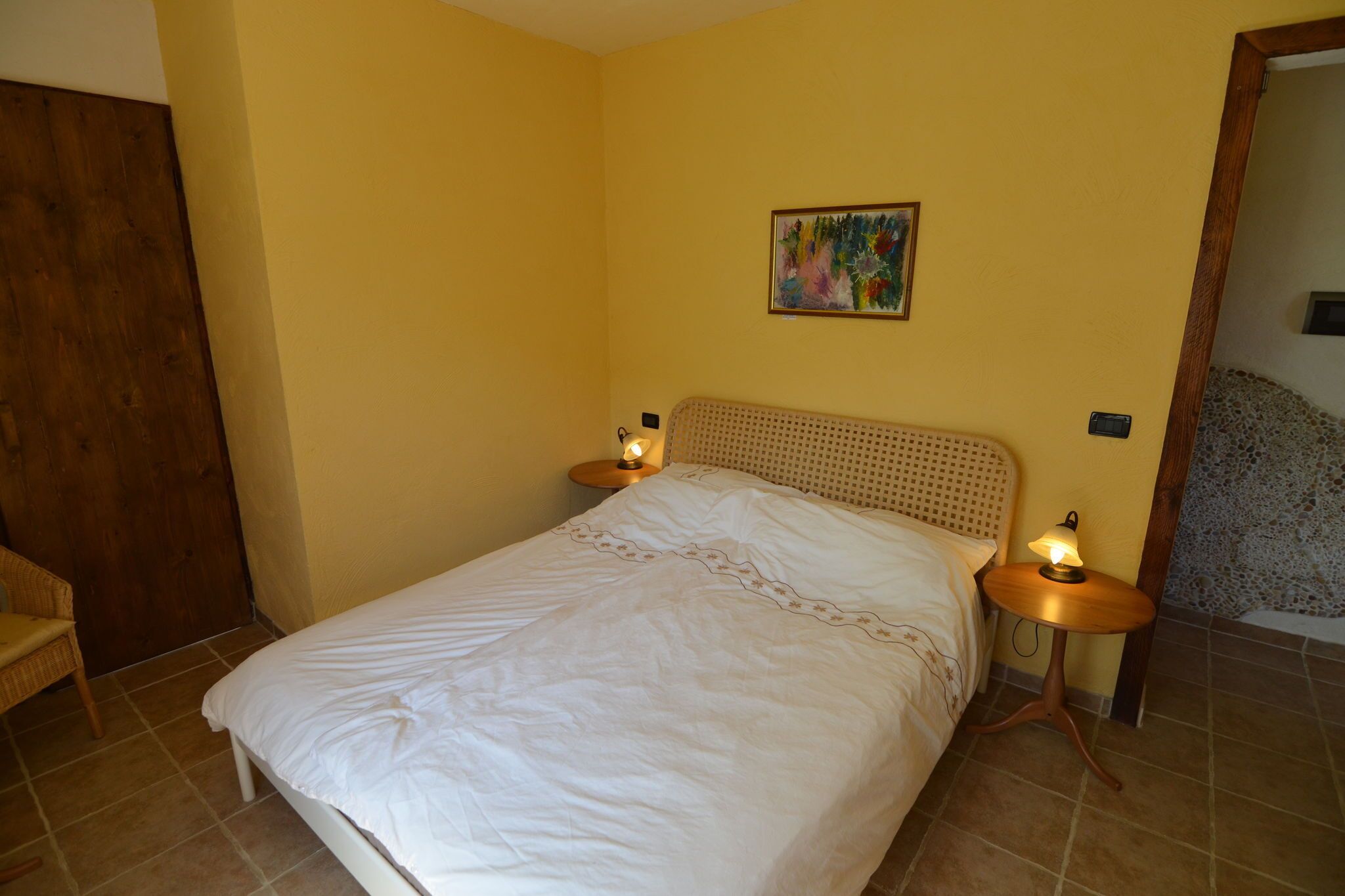 A bedroom with a bed, two bedside lamps, a wooden headboard, a painting on the wall, and a door.