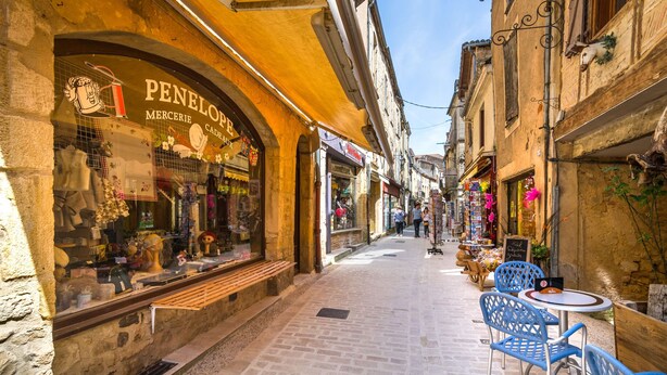 Una calle estrecha con tiendas, mesas al aire libre y edificios de piedra.