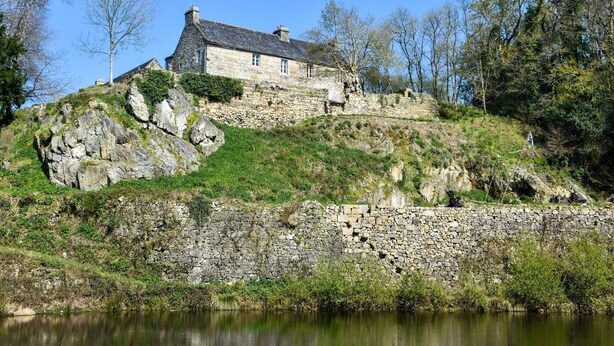 Une maison en pierre avec un toit en pente, située sur une colline surplombant une étendue d’eau.