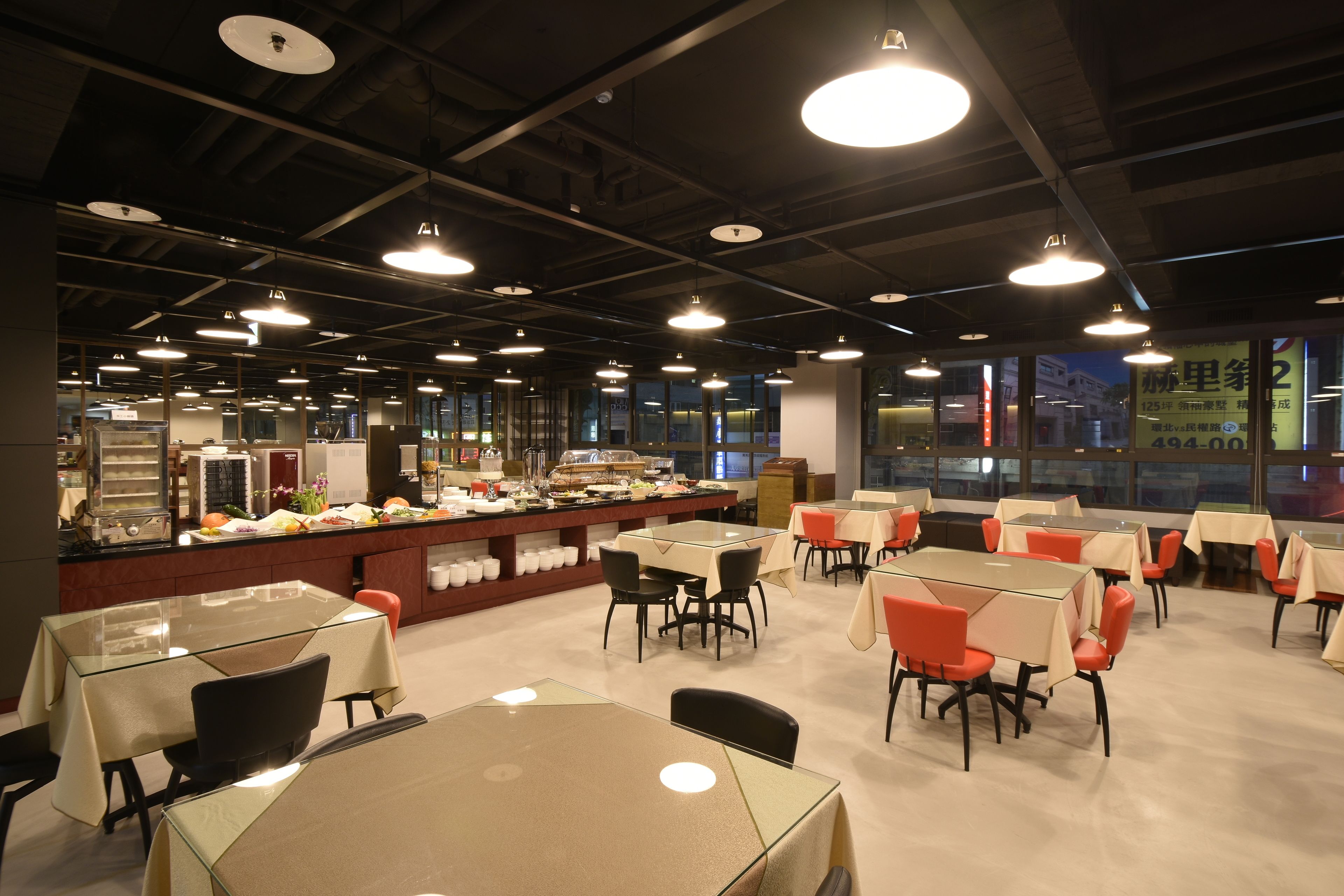 A spacious dining area with multiple tables and chairs, a central counter with food display, and large windows providing a view of the city.