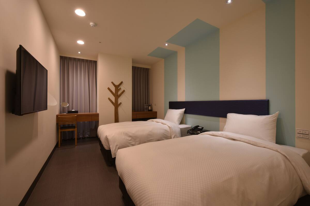 Una camera d'albergo con due letti, una scrivania, una sedia, una televisione e un appendiabiti.