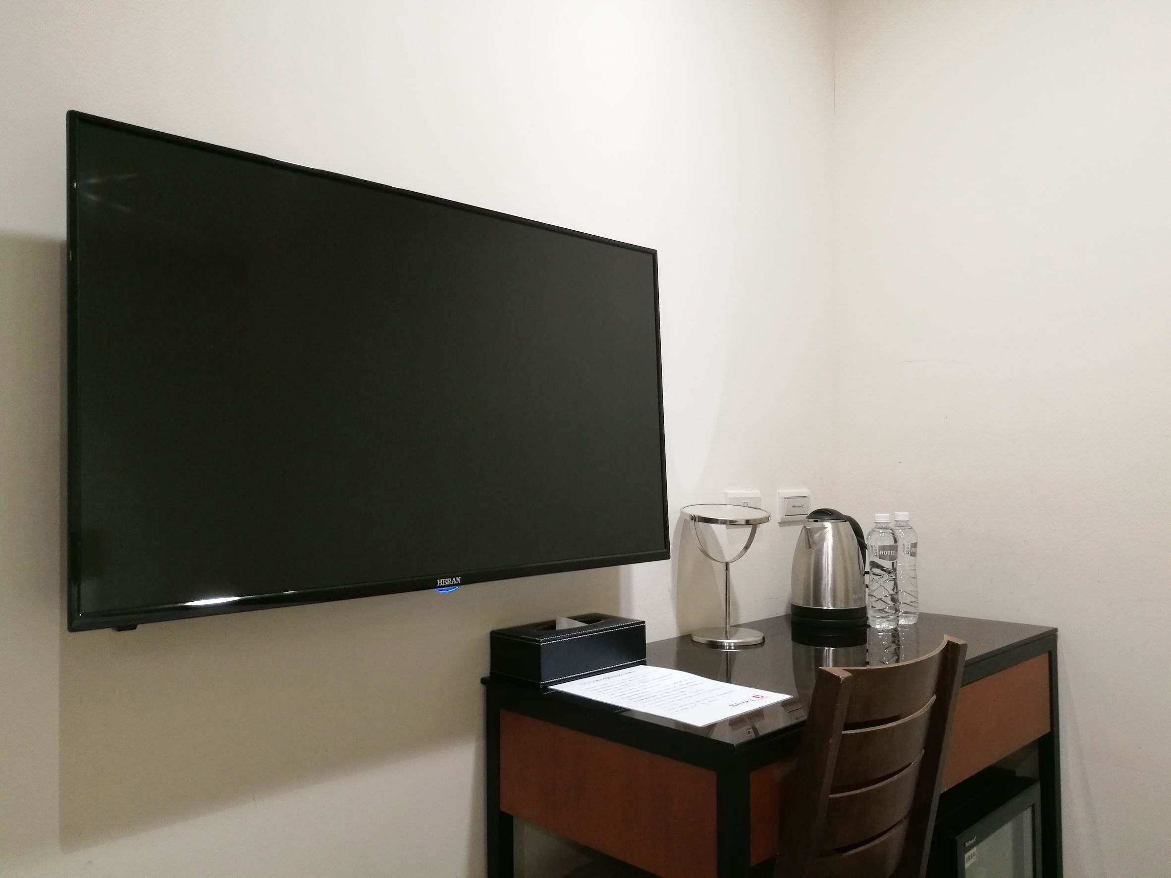 A hotel room with a flat-screen TV mounted on the wall, a desk with a notepad, a glass, a water bottle, and a coffee maker.