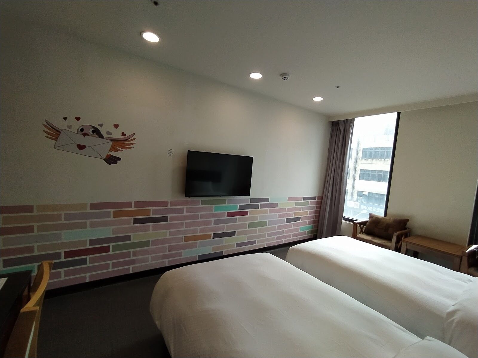 A hotel room with two beds, a flat-screen TV mounted on the wall, a brick-patterned accent wall, a window with a view of buildings, and a small table with a chair.