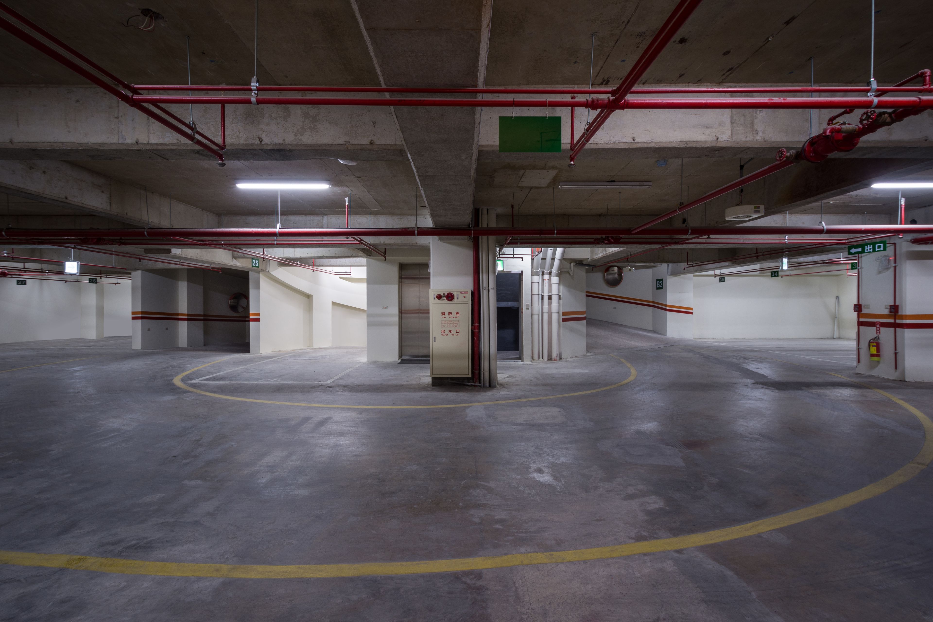 An indoor parking garage with concrete floors, red and white structural elements, and emergency exit signs.