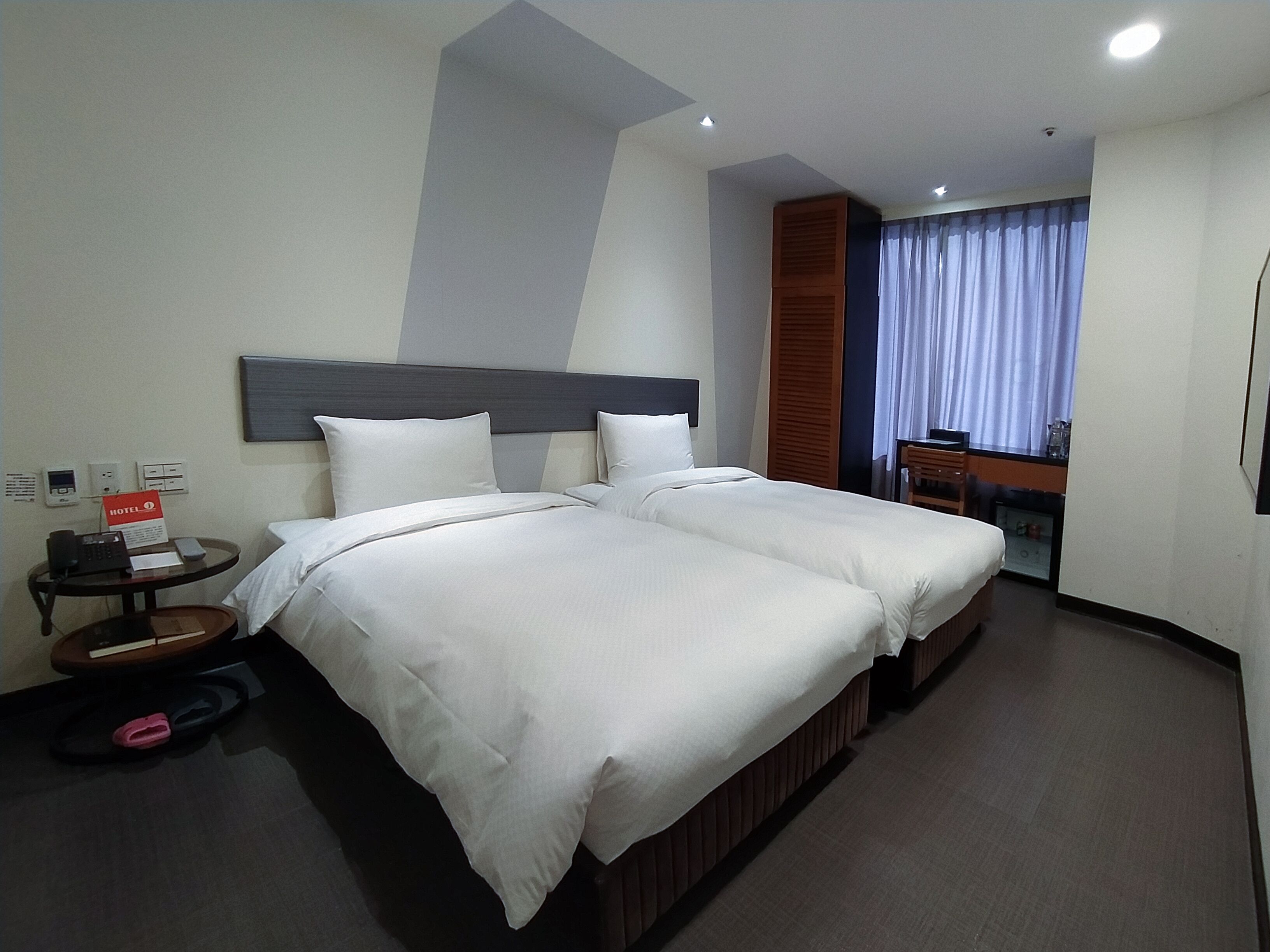 A hotel room with two beds, a desk, a chair, a small table, and a window with curtains.