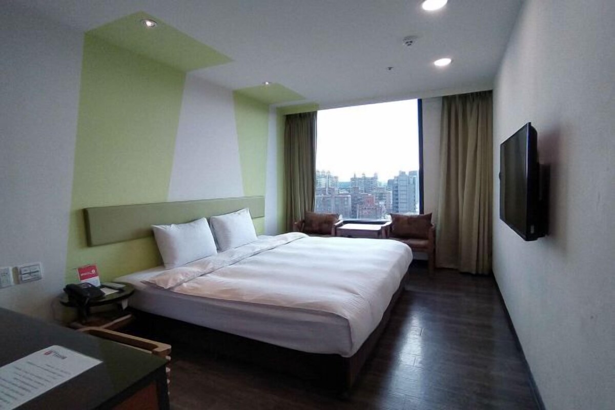 Ein Hotelzimmer mit einem großen Bett, einem Schreibtisch mit Telefon, einem an der Wand befestigten Fernseher und Blick auf die Stadt.