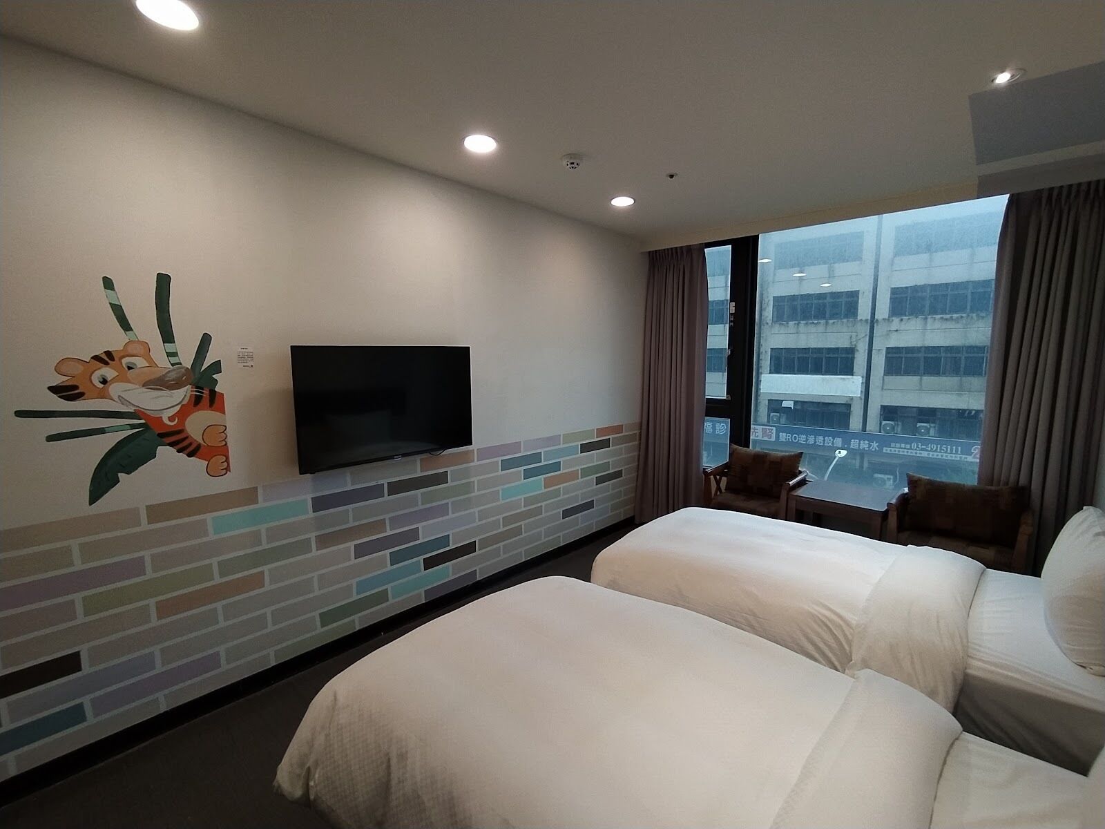 A hotel room with a large TV mounted on a brick-patterned wall, two beds, a window with a city view, and a small table with chairs.