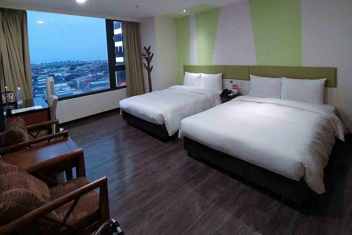Camera d'albergo con due letti, una scrivania, una sedia e una grande finestra con vista sulla città.