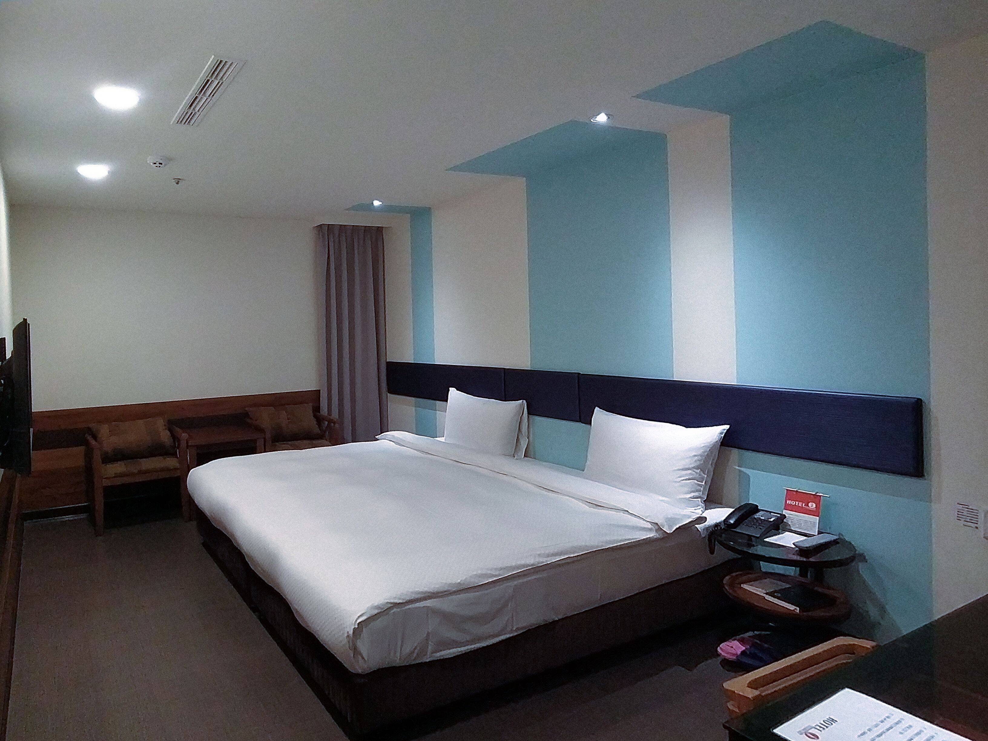 Habitación de hotel con cama, escritorio, silla y mesita de noche con teléfono y cartel.