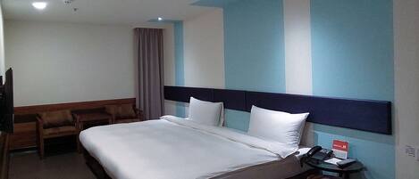 A hotel room with a bed, a desk, a chair, and a nightstand with a phone and a sign.