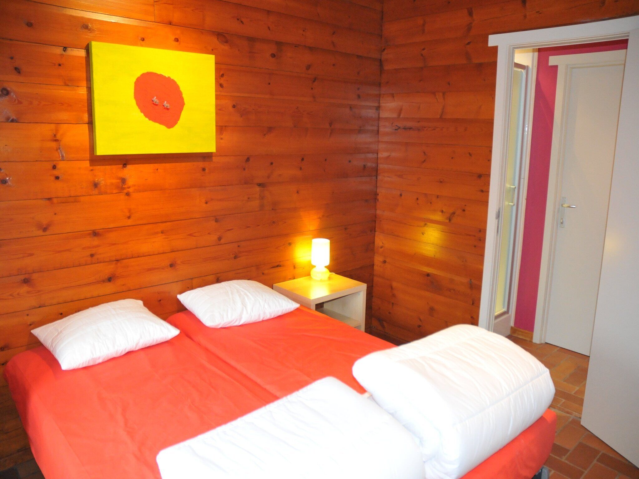 A room with wooden walls, a bed with orange bedding, white pillows, and a bedside table with a lamp.