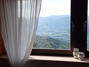 A room with a window overlooking a valley, a curtain, and a small table with decorative items. - Ferienhaus im Bayerischen Wald (Schöfweg ot Langfurth)