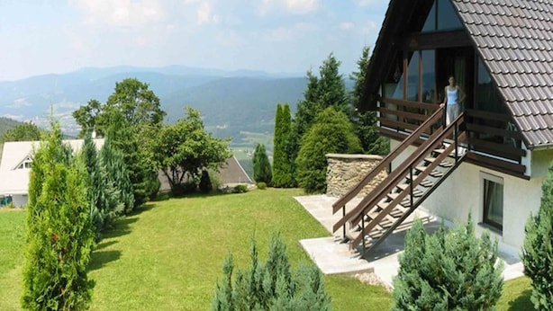 Ein modernes Haus mit einer Holztreppe, die auf einen Balkon führt, umgeben von einem gepflegten Rasen und Bäumen.