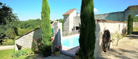 A stone building with a swimming pool, surrounded by tall cypress trees and a gravel path.