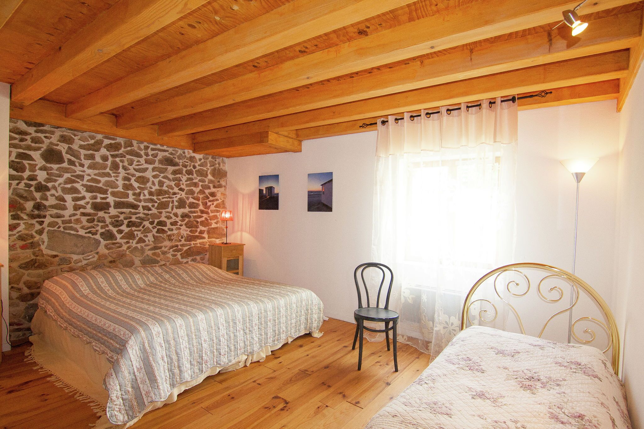 Un dormitorio con pared de piedra, vigas de madera en el techo, cama, silla y mesita de noche con lámpara.