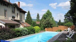 A swimming pool with a wooden deck and lounge chairs, surrounded by lush greenery and a house with a tiled roof.