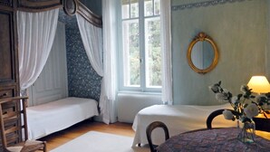 A room with two single beds, a wooden chair, a table with a vase of flowers, a mirror, and a window with curtains.