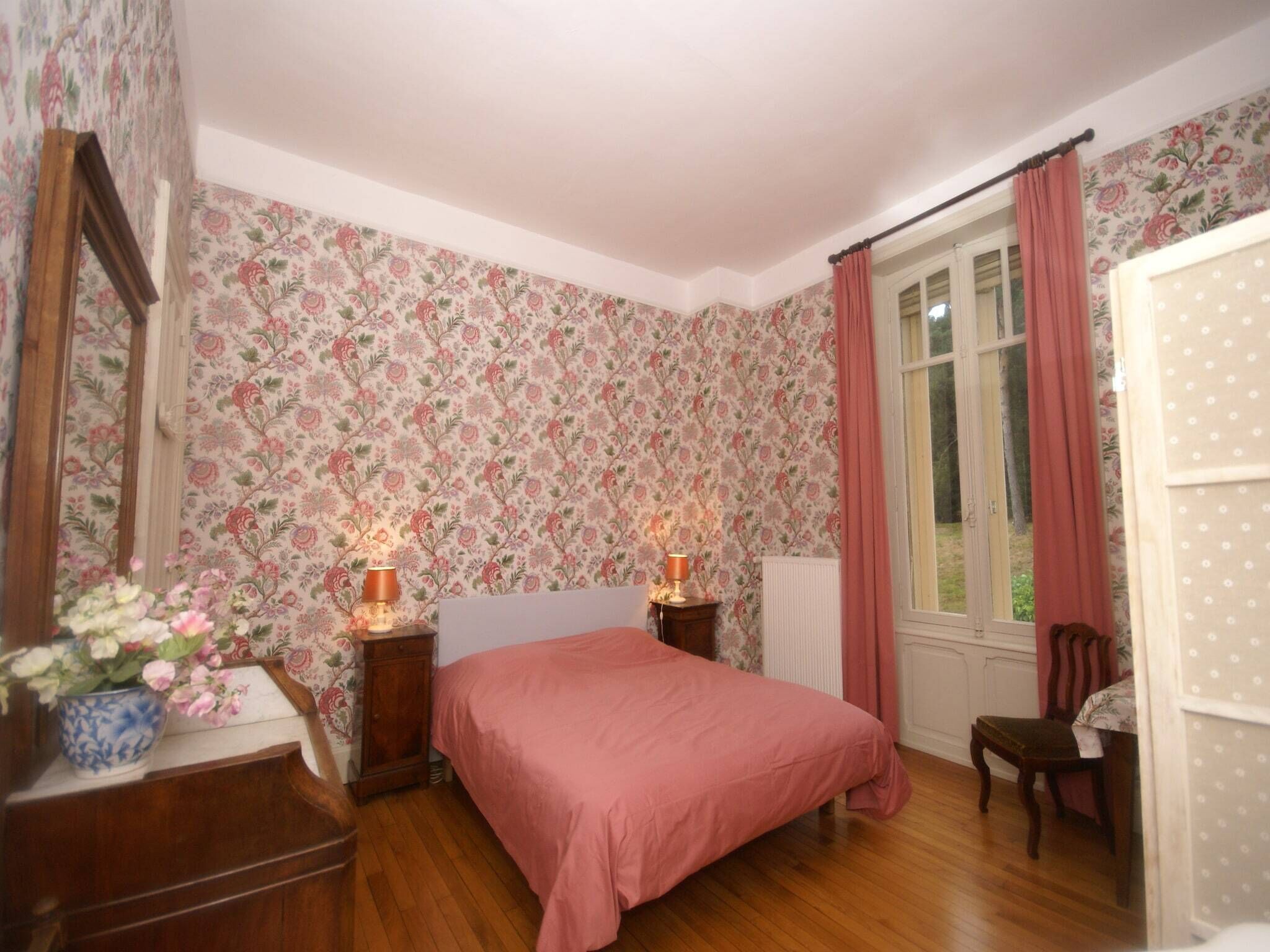 Un dormitorio con papel tapiz floral, una cama con colcha rosa, dos lámparas de noche, un aparador de madera y una ventana con cortinas.