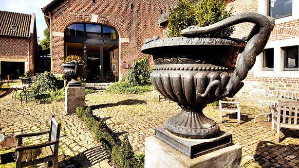 A courtyard with a large urn, outdoor seating, and a brick building with a glass entrance. - Pleasing Holiday Home in Richelle with Garden, Terrace, BBQ (Richelle)